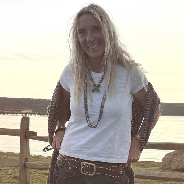 A woman posing outdoors on a grassy lawn near a wooden fence and water at sunset, wearing a white t-shirt, dark wide-leg jeans, and white sneakers.