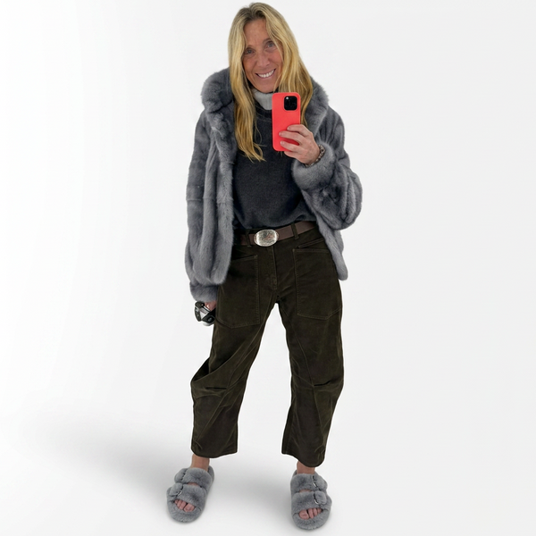 Person wearing a gray fur coat and dark pants, taking a selfie on a white background