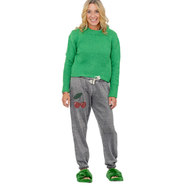 Woman wearing green fur slide sandals with a green sweater and gray pants with red cherry designs.