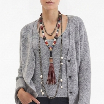 A model stands against a plain white studio background wearing a gray knit cardigan, dark navy wide-leg trousers, layered necklaces, and black fuzzy slide sandals.