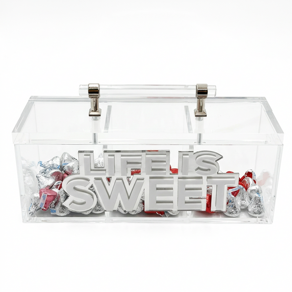 A clear acrylic candy box with a handle, filled with silver foil wrapped chocolates and featuring the words 'LIFE IS SWEET' in large 3D letters on the front.