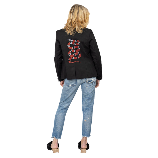 Full-length back view of a woman wearing a black blazer with a red and silver rhinestone snake, blue jeans and black heels.