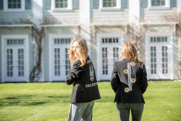 Two women outdoors showing black blazers with a skull and lettering design and a floral design.